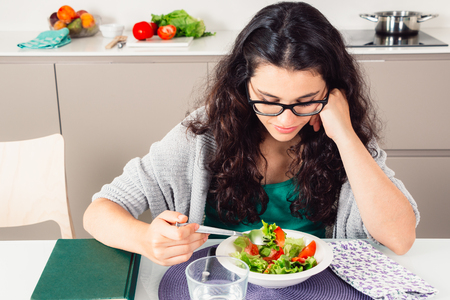 Young woman with glasses eating a boring salad in the kitchen with a book next to herの写真素材