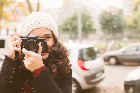 Young beautiful woman with a woolen cap taking pictures with a retro camera in the city in autumn....