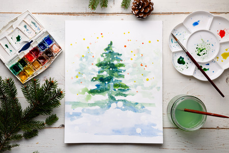 Handmade watercolor Christmas card on the work table with watercolor box, brushes, palette and fir branches and pine cones. Top viewの写真素材