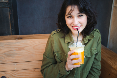Young happy woman with green jacket drinking a fruit smoothie with a straw outdoorsの写真素材