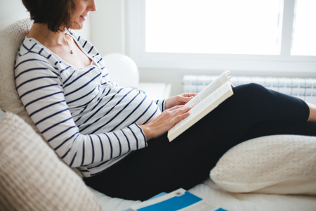 Happy pregnant woman in striped t-shirt sitting on the bed with legs on a cushion reading books about pregnancyの写真素材