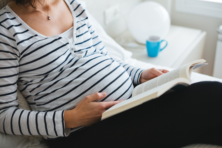 Happy pregnant woman in striped t-shirt sitting on the bed with legs on a cushion reading books about pregnancy, close upの写真素材