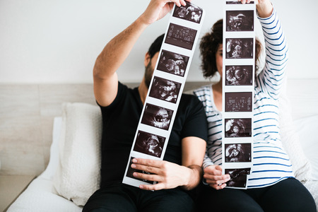 Happy and excited pregnant couple showing stripes of ultrasound images of their baby sitting on the...
