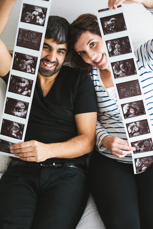 Happy and excited pregnant couple showing stripes of ultrasound images of their baby sitting on the bedの写真素材
