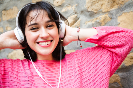 Happy young girl in pink sweater listening to music in white headphones leaning in a street wall.の写真素材