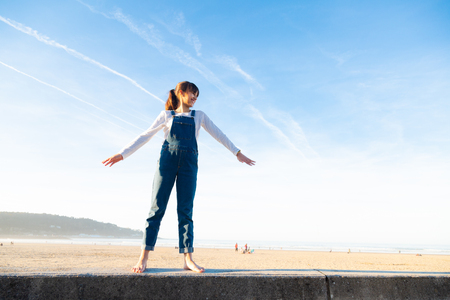 Happy little girl in overalls raising arms like a plane against the sky on the beachの写真素材
