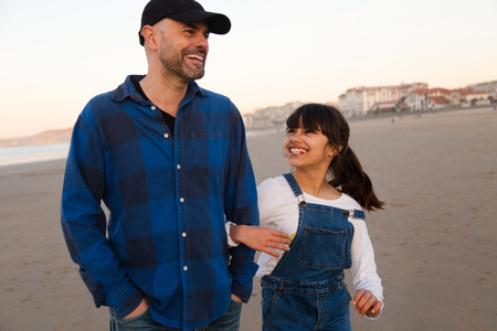 Funny father and daughter laughing while walking arm in arm on the beach at sunsetの写真素材