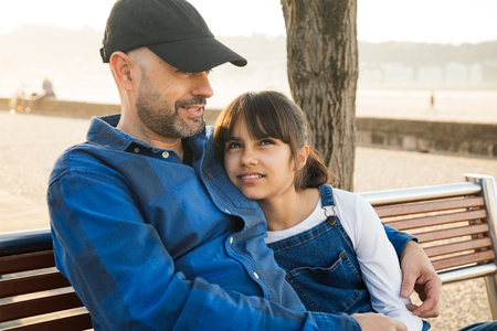 Father and daughter sitting on a bench on the waterfront having a good conversation in a sunny dayの写真素材