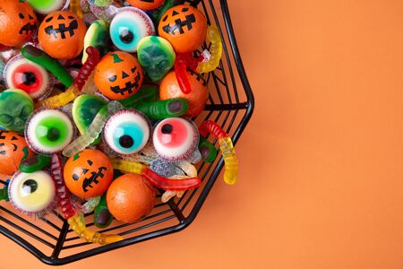 Spiderweb shaped bowl full of candy and chocolate with shape of eyes, worms, fingers, spiders and monsters on an orange background. Top viewの写真素材