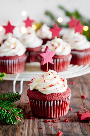 Christmas red velvet cupcakes decorated with a star shaped cake pick, with Christmas ornaments, fir and lights on a rustic wooden tableの写真素材