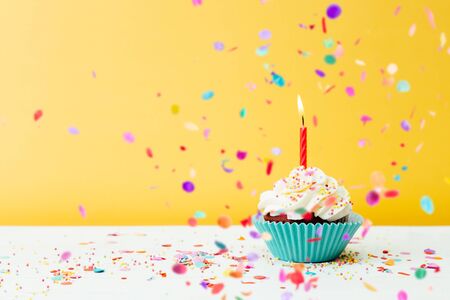 A colorful  birthday cupcake with one candle and confetti on a yellow backgroundの写真素材