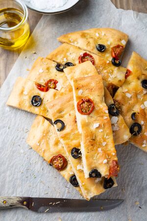 Homemade focaccia with black olives, cherry tomatoes, rosemary, flake salt and olive oil on baking paper. Top viewの写真素材