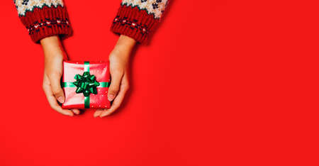 Female hands with a winter sweater holding a Christmas gift on a red background. Copy spaceの写真素材