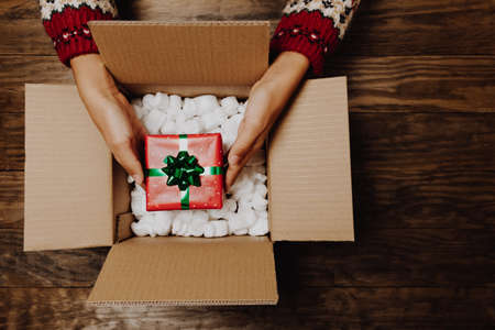 Female hands with a winter sweater packing or unpacking a Christmas gift from a cardboard box on a rustic wooden table. Christmas home delivery or shipping concept. Top viewの写真素材