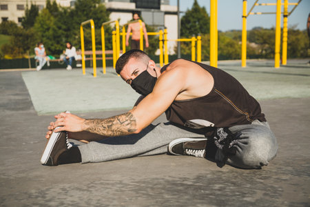 Athletic young man with black mask warm up by stretching legs before exercises at the calisthenics...