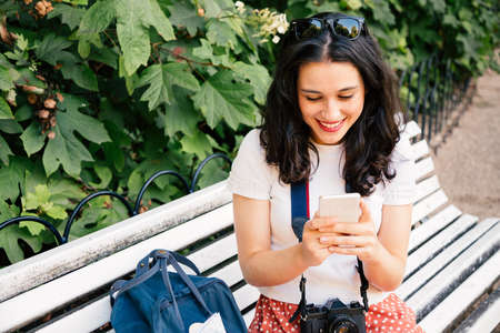 Happy tourist woman using her martphone sitting on a park bench. Addicted to technologyの写真素材
