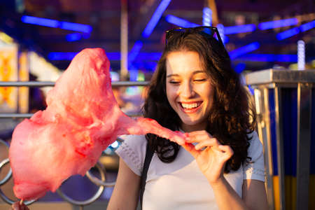 Happy young beautiful woman eating cotton candy at fairground at sunsetの写真素材