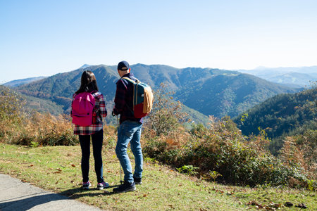 Father and his teenage daughter with backpacks and sticks hiking in autumn looking at the landscape in a sunny day. Back viewの写真素材
