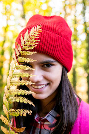 Smiling teen girl with a red woolen cap and pink backpack covering face with a dry fern in autumn in the forestの写真素材