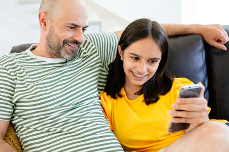 Happy father and teen daughter watching something in her smart phone or having a video call sitting in the sofaの写真素材