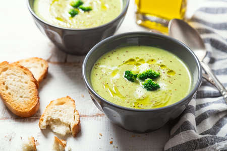 Bowls of broccoli cream soup with olive oil, pepper and toasted bread slices on a rustic white wooden tableの写真素材