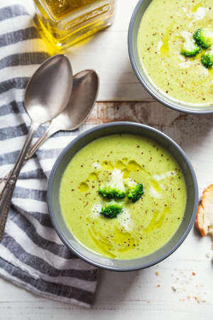 Bowls of broccoli cream soup with olive oil, pepper and toasted bread slices on a rustic white wooden tableの写真素材
