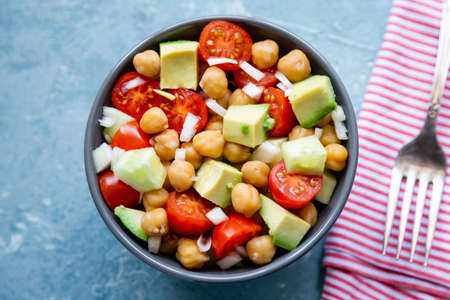 Healthy and delicious chickpea salad with  cherry tomato, avocado, cucumber and onion in a bowl un a rough blue backgroundの写真素材