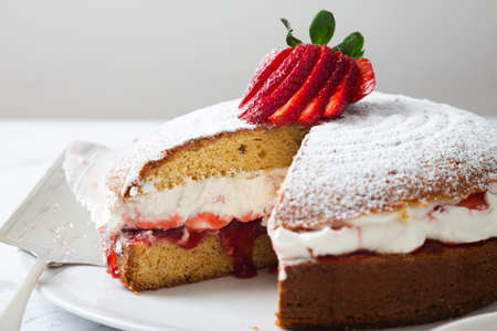 Homemade Victoria sponge cake filled with strawberries, jam and whipped cream decorated with icing sugar and strawberry on a white marble tableの写真素材