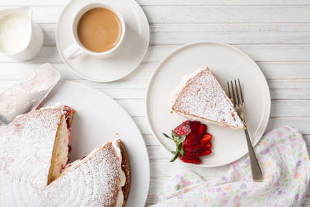 Top view of homemade Victoria sponge cake filled with strawberries, jam and whipped cream  on a white wooden table and a cup of coffee or teaの写真素材