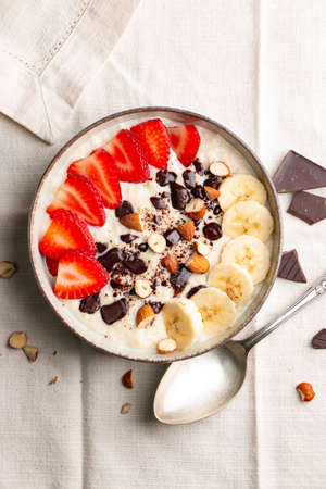 Millet porridge with strawberry, banana, dark chocolate pieces, hazelnut and almond in a ceramic bowl on a white napkinの写真素材