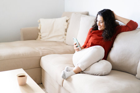 Young happy woman sitting comfortably on the couch using her smart phone and having a beverageの写真素材