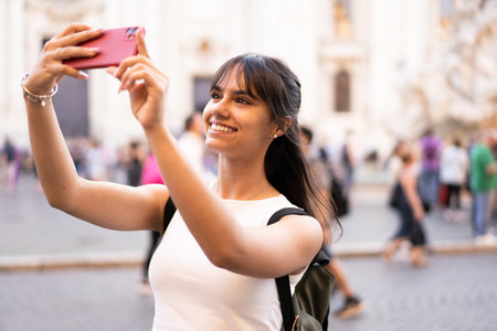 Happy teen girl with backpack taking a selfie In a crowded square in Rome on vacationの写真素材