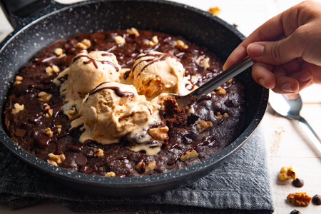 Homemade brownie cooked in a skillet, not baked, with melting vanilla ice cream scoops, nuts, and chocolate chips and a woman's hand taking a piece with a spoonの写真素材