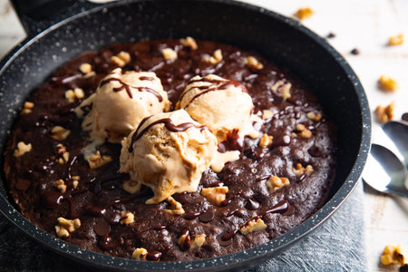 Homemade brownie cooked in a skillet, not baked, with melting vanilla ice cream scoops, nuts, and chocolate chips on a white tableの写真素材