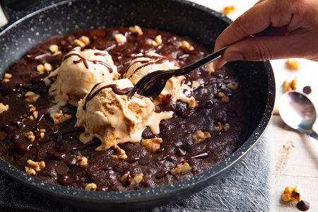 Homemade brownie cooked in a skillet, not baked, with melting vanilla ice cream scoops, nuts, and chocolate chips and a woman's hand taking a piece with a spoonの写真素材