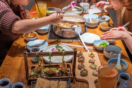 Table full of dishes and ingredients to be cooked in hotpot restaurant, with three dinersのeditorial素材