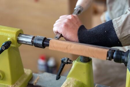 Carpenter man is carving the wooden stick. Spinning wood wheel machine. Trimming forming a round detail. Joiner is using metal tool to make the handmade decoration at workshop.の写真素材