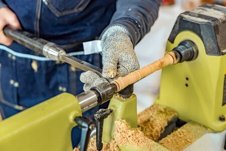 Carpenter man is carving the wooden stick. Spinning wood wheel machine. Trimming forming a round detail. Joiner is using metal tool to make the handmade decoration at workshop.の写真素材