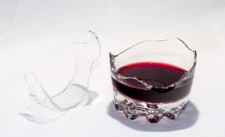 A broken whiskey glass with the remains of red wine stands on a white background. There are splinters nearby.の写真素材