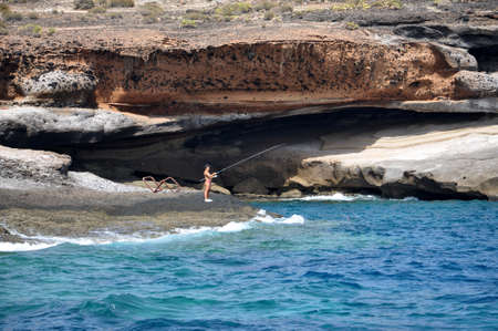 TENERIFE, SPAIN - JULY 1, 2011: Unknown man standing on the beach of ocean and with fishing rod catch fish.の写真素材