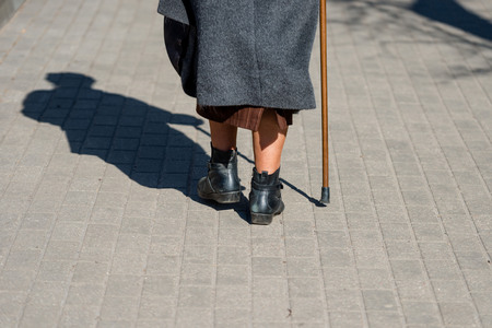 On a sunny day a old woman walking down the street with walking stick. The woman's shadow is visible on the sidewalk. View from the back.の写真素材