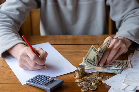 Close up man with calculator counting, making notes at home, hand is writes in a notebook. Stacked coins arranged at deesk. Savings finances concept.の写真素材