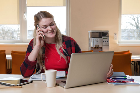 A smiling blonde woman sitting at a laptop and talking over a cell phone.の写真素材