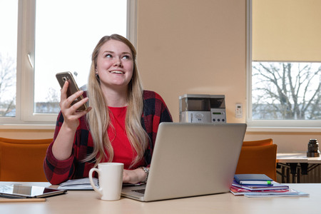 A smiling blonde woman sitting at a laptop and talking over a cell phone.の写真素材
