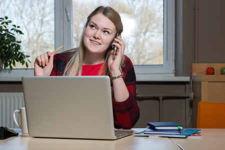 A smiling blonde woman sitting at a laptop and talking over a cell phone.の写真素材