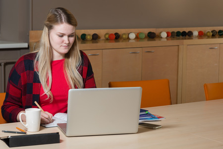 Smiling blonde woman sitting at a laptop drinking tea and working.の写真素材