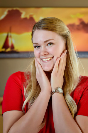 Surprised smiling blonde woman with long blonde hair and hands on face in a red dress.の写真素材