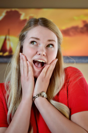 Surprised smiling blonde woman with long blonde hair and hands on face in a red dress.の写真素材