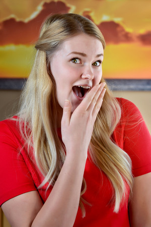 urprised and excited blonde woman with long blonde hair and hands on face in a red dress.の写真素材