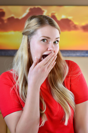 urprised and excited blonde woman with long blonde hair and hands on face in a red dress.の写真素材